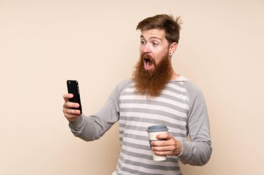 Uzun sakallı kızıl saçlı bir adam izole edilmiş bir arka planı var. Elinde kahve ve cep telefonu var.