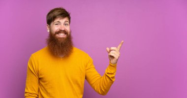 Uzun sakallı kızıl saçlı adam, izole edilmiş mor arka planda, parmağını yana doğru gösteriyor.