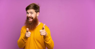 Uzun sakallı, soyutlanmış mor arka planı olan kızıl saçlı adam ön tarafı işaret ediyor ve gülümsüyor.
