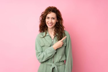 Soyutlanmış pembe arka planda genç ve güzel bir kadın bir ürünü sunmak için kenarı işaret ediyor.