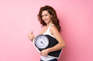 Kıvırcık saçlı, tartı makinesini soyutlanmış pembe arka planda tutan genç bir kadın.