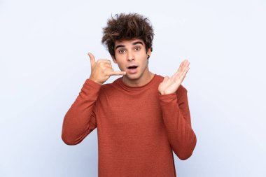 İzole edilmiş mavi arka planda genç beyaz bir adam telefon jesti yapıyor ve şüpheli.