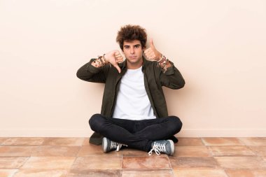 Genç, beyaz bir adam yerde oturmuş iyiye-kötüye işaret veriyor. Evet ya da hayır arasında kararsız