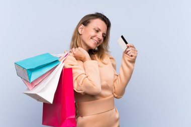 Sarışın kadın izole edilmiş mavi arka planda elinde alışveriş çantaları ve kredi kartı tutuyor.