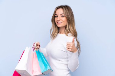 Soyutlanmış mavi arka planda genç bir kadın elinde alışveriş torbalarıyla ve başparmağıyla