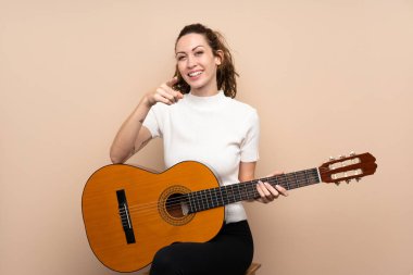 Arka planda gitar çalan genç bir kadın kendinden emin bir ifadeyle seni işaret ediyor.