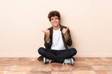 Genç, beyaz bir adam yerde oturuyor. Başparmakları havada ve gülümsüyor.