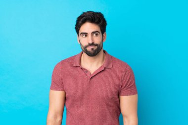 Mavi arka planda sakallı yakışıklı genç adam şüpheli hareketler yapıyor.