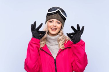 Snowboard gözlüklü genç kız izole edilmiş mavi arka planda parmaklarıyla iyi bir işaret gösteriyor.