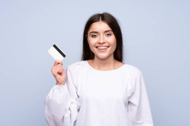 Soyutlanmış mavi arka planı olan genç bir kadın kredi kartı tutuyor.