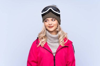 Snowboard gözlüklü kayakçı kız mavi arka planda duruyor ve yan tarafa bakıyor.