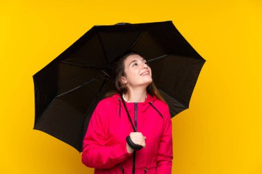 Sarı arka planda şemsiyesi olan esmer kız gülümserken yukarı bakıyor.