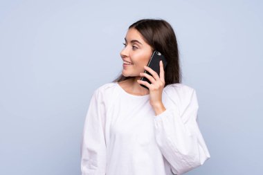 İzole edilmiş mavi arka planı olan genç bir kadın cep telefonuyla sohbet ediyor.