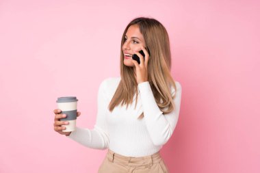 Soyutlanmış pembe arka planı olan genç sarışın bir kadın elinde kahve ve bir cep telefonu tutuyor.