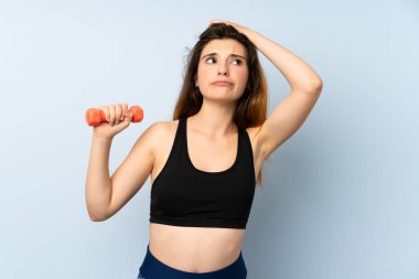 Yalıtılmış mavi arka planda ağırlık kaldıran genç spor kadınının şüpheleri ve kafa karıştırıcı bir yüz ifadesi var.