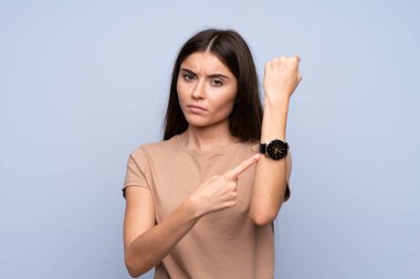 İzole edilmiş mavi arka planda genç bir kadın el saatini ciddi bir ifadeyle gösteriyor çünkü geç oluyor.