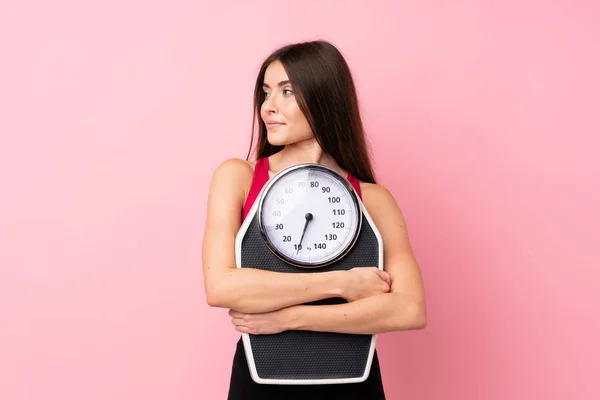 Pretty young girl with weighing machine over isolated pink background with weighing machine and looking side