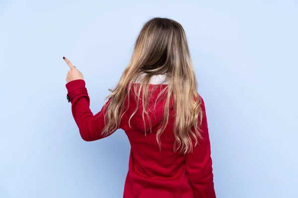 穿着冬衣的年轻女人用食指指向后方的孤立的蓝色背景 — 图库照片