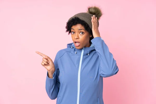 アフリカ系アメリカ人女性とともに冬の帽子以上隔離されたピンクの背景驚きと指差し指へ側 — ストック写真
