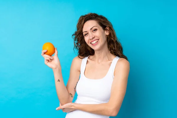 Ung kvinna med lockigt hår håller en orange över isolerad blå bakgrund — Stockfoto