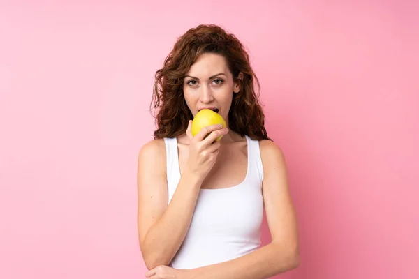 Ung kvinna med lockigt hår håller ett äpple över isolerad rosa bakgrund — Stockfoto