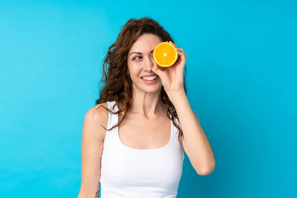 Ung kvinna med lockigt hår håller en orange över isolerad blå bakgrund — Stockfoto