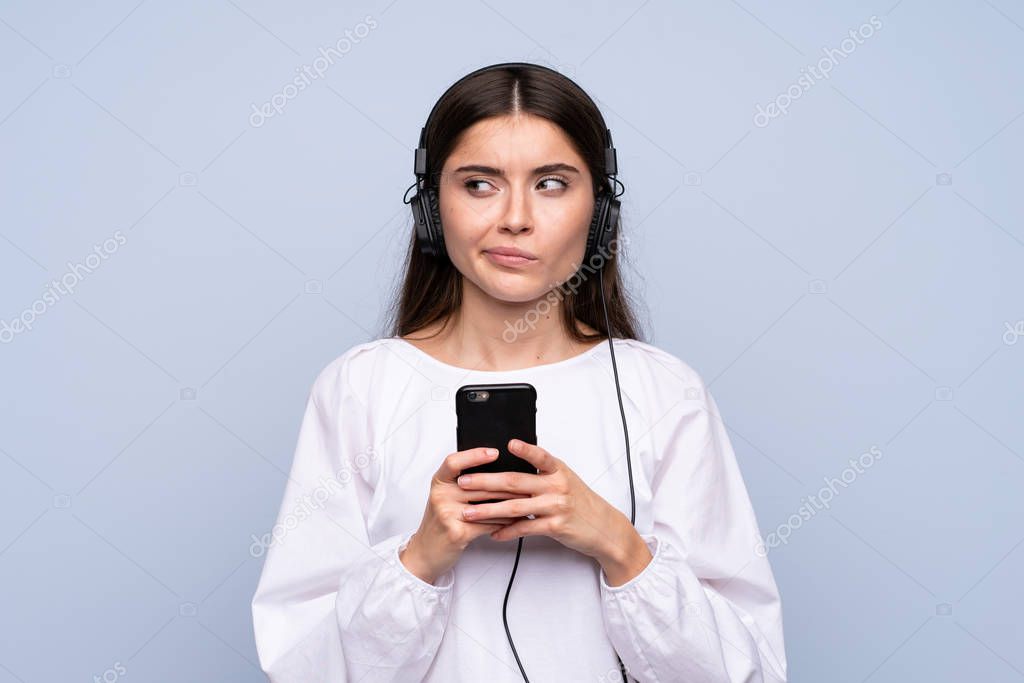 Young woman over isolated blue background using the mobile with headphones and thinking