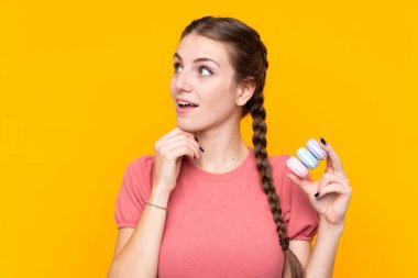 Young woman over isolated yellow background holding colorful French macarons and thinking an idea