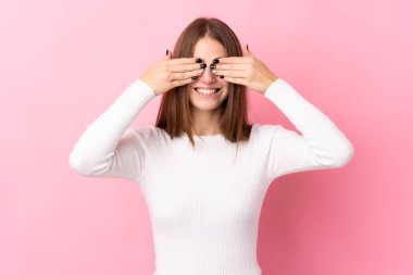 İzole edilmiş pembe arka planı olan genç bir kadın elleriyle gözlerini kapatıyor.