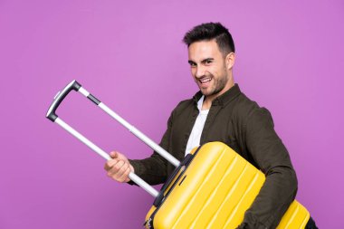 Young handsome man over isolated purple background in vacation holding a travel suitcase like a guitar