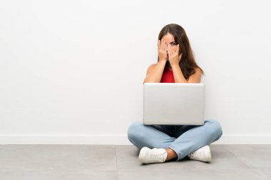 Dizüstü bilgisayarı olan genç bir kadın yerde oturuyor gözleri kapatıyor ve parmaklarından bakıyor.