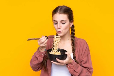 İzole edilmiş sarı arka plan üzerinde genç bir kadın elinde çubuklu bir kase erişteyle sıcak olduğu için üflüyor.