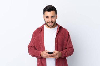 Sakallı, kadife ceket giyen genç yakışıklı bir adam izole edilmiş beyaz arka planda cep telefonuyla bir mesaj gönderiyor.
