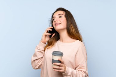 Genç esmer kız izole edilmiş mavi arka planda elinde kahve ve bir cep telefonu var.