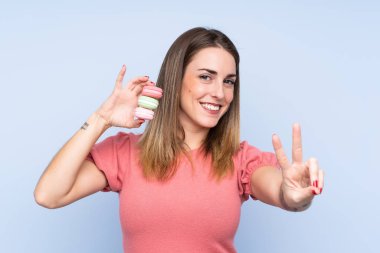 Soyutlanmış mavi arka planda genç sarışın kadın elinde renkli Fransız kurabiyeleri ve zafer işareti taşıyor.