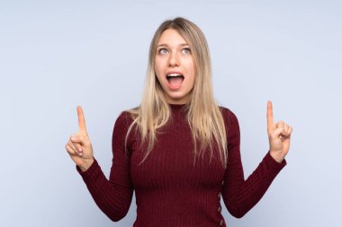 İzole edilmiş mavi arka planı olan genç sarışın kadın şaşırmış ve yukarı bakıyor.