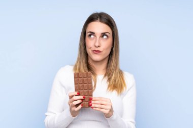 Genç sarışın kadın, izole edilmiş mavi arka planda çikolata tableti alıyor ve kuşkuları var.