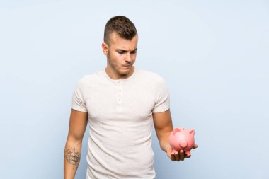 Soyutlanmış mavi arka planı olan yakışıklı sarışın adam elinde büyük bir kumbara tutuyor.