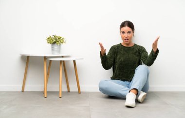 Genç bir kadın yerde oturuyor ve yüzünde sürpriz bir ifade var.