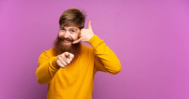 Uzun sakallı, uzun boylu, mor arkaplanı olan, telefon hareketleriyle ön tarafı işaret eden kızıl saçlı bir adam.