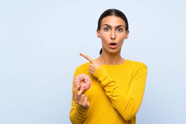 Mavi arka planda donut tutan genç bir kadın şaşırmış ve işaret ediyor.