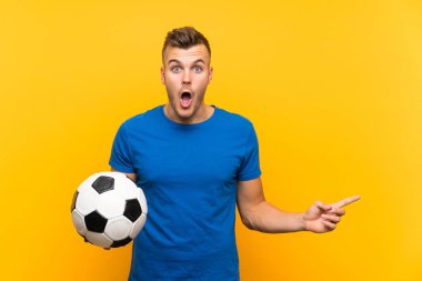Sarı arka plan üzerinde futbol topu tutan yakışıklı sarışın adam şaşırmış ve işaret ediyor.