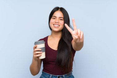 Genç Asyalı kız elinde kahveyle geziyor gülümsüyor ve zafer işareti gösteriyor