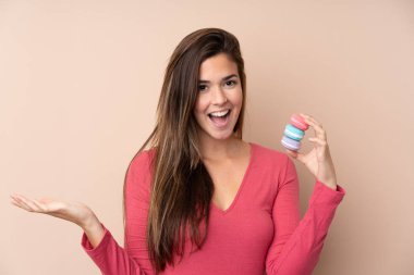 Soyutlanmış arka planda genç bir kız elinde renkli Fransız kurabiyeleriyle şok olmuş bir ifade var.