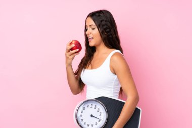 Soyutlanmış pembe arka plan, tartı makinesi ve elması olan genç bir kadın.