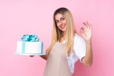 Genç Uruguaylı pastacı kadın, izole edilmiş pembe arka planda büyük bir pastayla parmaklarıyla tamam işareti gösteriyor.