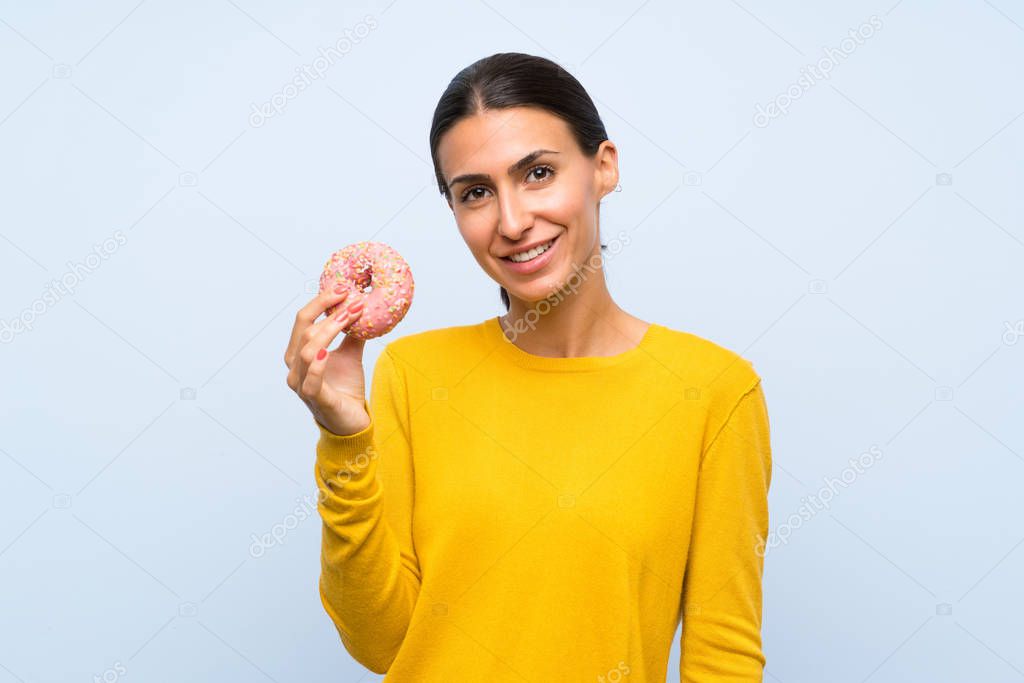 Young woman holding a donut over isolated blue background smiling a lot
