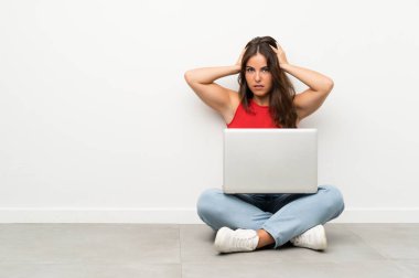 Dizüstü bilgisayarı olan genç bir kadın yerde oturuyor ve yüzünde sürpriz bir ifade var.