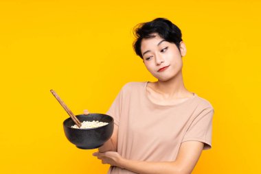 Asyalı genç bir kız, yemek çubuklarıyla bir kase erişte tutarken üzgün bir ifadeyle izole edilmiş sarı arka planda.