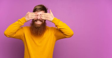 Uzun sakallı kızıl saçlı adam, tek başına mor arka planda, gözleri elleriyle kapatıyor.